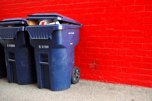 Two large dark blue plastic wheelie bins with black lids are positioned on a concrete pavement next to a red brick wall. The bins are marked with white labels, and one bin's lid is slightly open, revealing some mixed rubbish inside. The bins are situated on the left side of the image, with the background showing a clean, well-maintained red brick wall that extends horizontally across the scene. The overall environment is outdoors, likely on a residential or commercial property driveway or alleyway in Newham, where rubbish collection and waste management services, such as those provided by Commercial Waste Newham, are commonly scheduled to handle waste disposal efficiently.