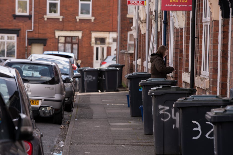 A row of five large black wheeled rubbish bins with yellow lids standing side by side on a concrete surface against a plain, light-colored wall. Each bin has a white label with printed numbers and tex