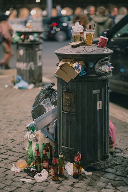 In the foreground, there is a large pile of mixed waste and rubbish consisting of plastic bags, cardboard, paper, broken furniture, and discarded household items, spread across a patch of ground with